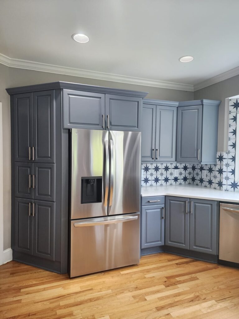 A modern kitchen with stainless steel refrigerator, blue-gray cabinets, white countertops, and a backsplash featuring a blue starburst tile pattern, set on a light wooden floor.
