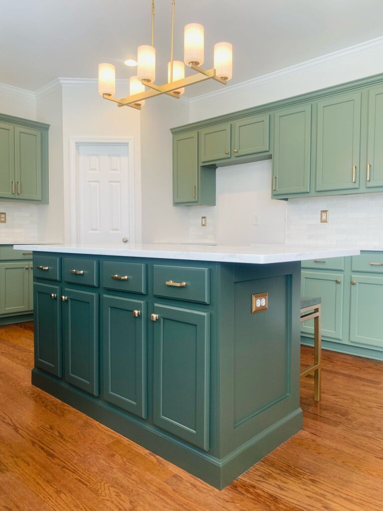 Modern kitchen with light green cabinets, a dark green island featuring gold hardware from expert cabinet painting Charlotte, white countertops, white tile backsplash, wood floors, and a gold chandelier with five lights.