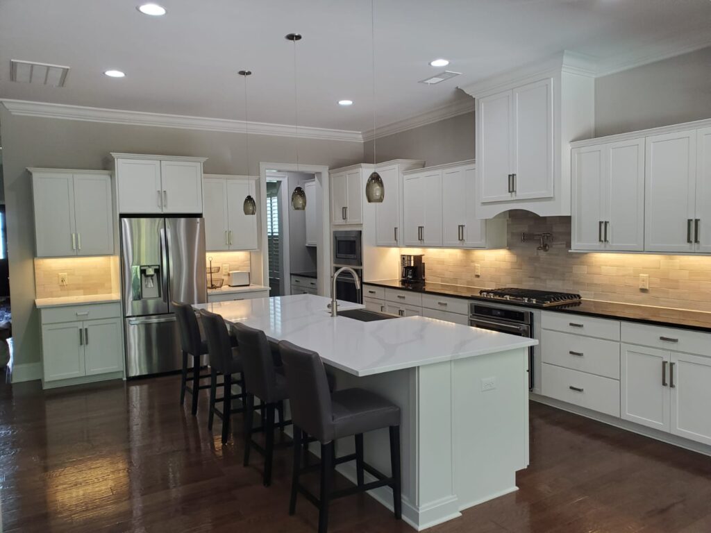 Modern kitchen featuring freshly finished white cabinets—showcasing expert cabinet painting Charlotte services—stainless steel appliances, a large island with four gray barstools, pendant lights overhead, and bright under-cabinet lighting.