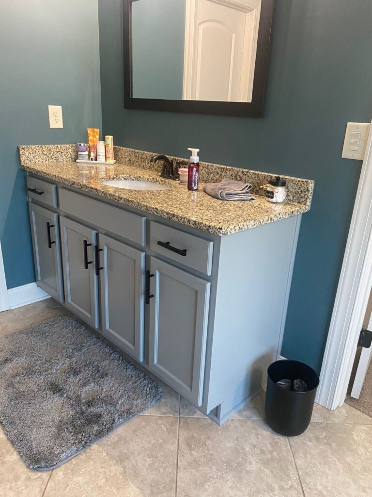 A bathroom vanity with a granite countertop, sink, soap dispenser, toiletries, folded towel, and cup. Light gray cabinets with black handles—showcasing quality cabinet painting Charlotte—gray rug on tiled floor, black trash can, and a large mirror above.