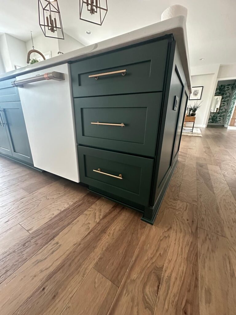 A kitchen island with dark green cabinets, finished by expert cabinet painting Charlotte, and gold handles, featuring a white dishwasher panel, sits on a light wood floor in a bright, modern kitchen.