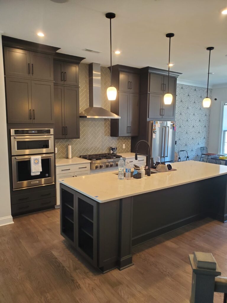 Modern kitchen with dark cabinets, cabinet painting Charlotte finishes, stainless steel appliances, a large island with a beige countertop, pendant lights above, herringbone backsplash, and geometric wallpaper in the background. Bottles and items sit on the island.
