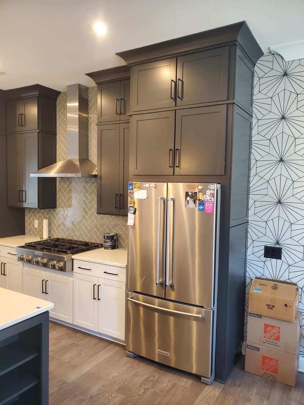 Modern kitchen with dark gray upper cabinets, white lower cabinets, a stainless steel refrigerator with magnets, a gas stove and hood, herringbone backsplash, and a Home Depot moving box—perfect for showcasing cabinet painting Charlotte style.