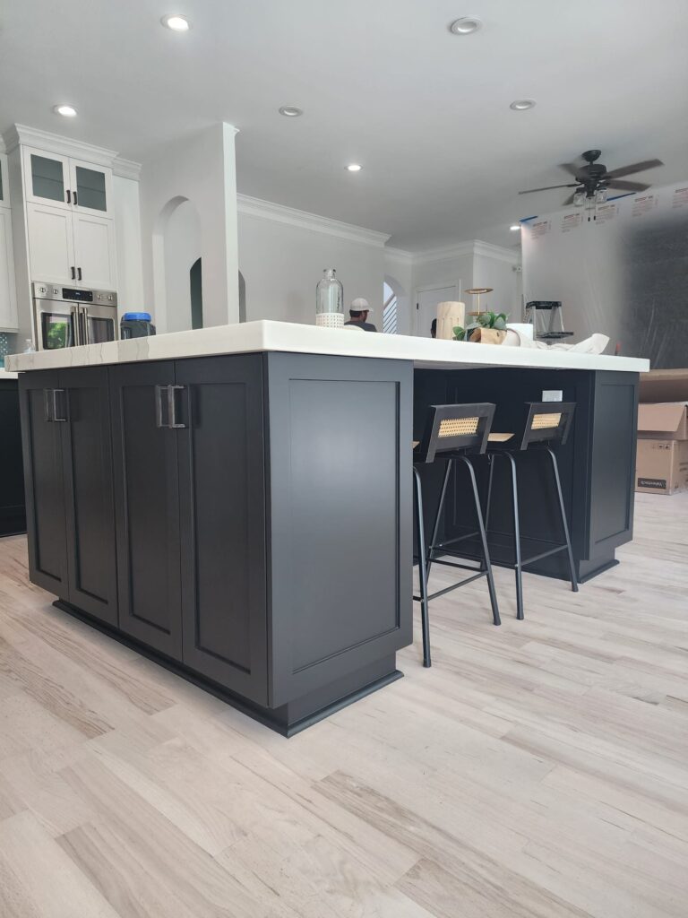 A modern kitchen with a large black island, white countertop, two black stools with woven seats, light wood flooring, and freshly painted white cabinets in the background—showcasing expert cabinet painting Charlotte. The space appears bright and open.
