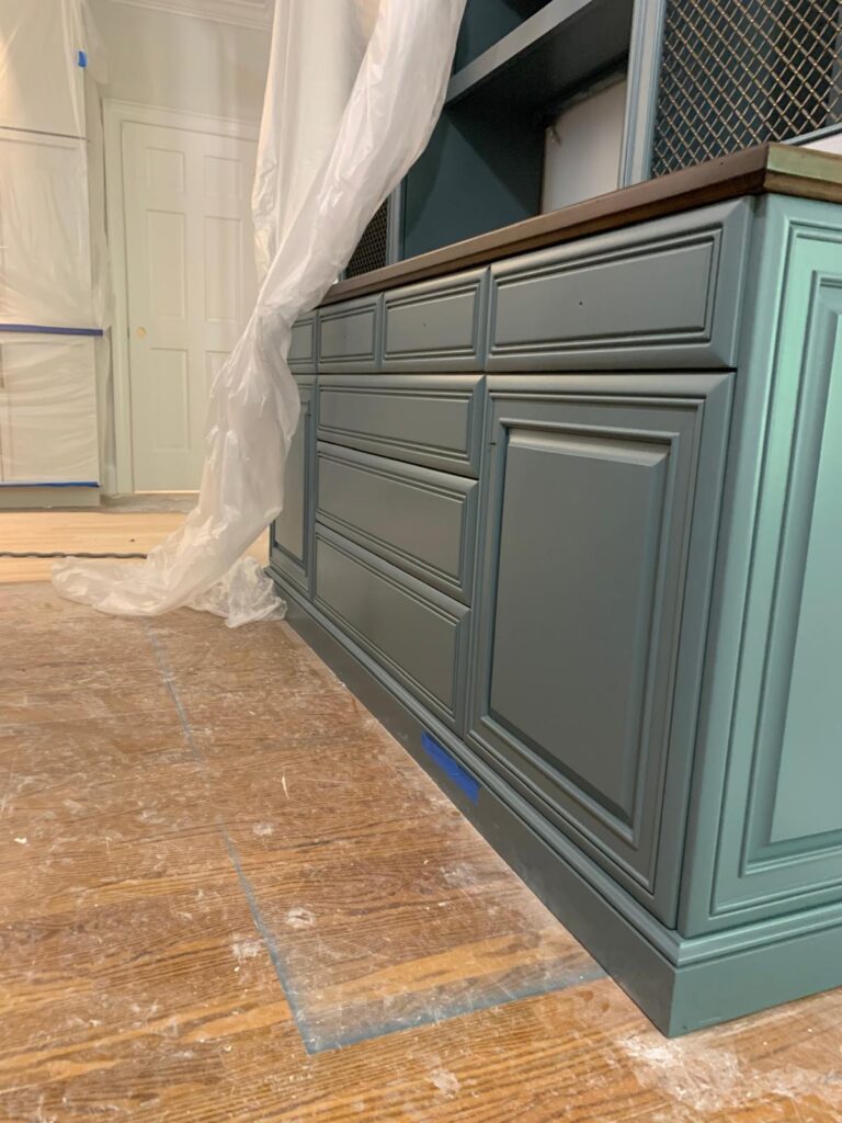 A close-up view of teal cabinets and drawers in a room under renovation, showcasing expert cabinet painting Charlotte, with plastic sheets draped above, painter’s tape on the floor, and protective covering on the wooden floor.