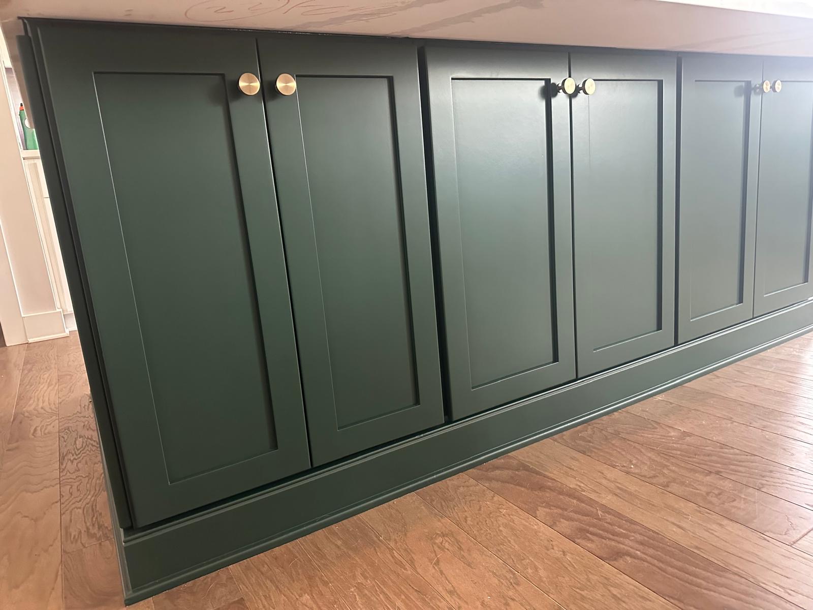 Dark green kitchen cabinets with simple, recessed panel doors and round brass knobs, set against a light brown wooden floor. The modern and clean design showcases the quality of cabinet painting Charlotte homeowners prefer.