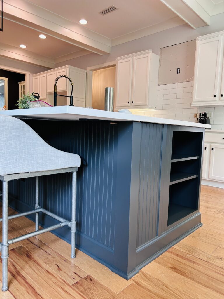 A modern kitchen features a navy blue island with a white countertop, open shelves, and a gray upholstered bar stool. Cabinet painting Charlotte gives the white cabinets a fresh look, complemented by wood flooring and a subway tile backsplash.