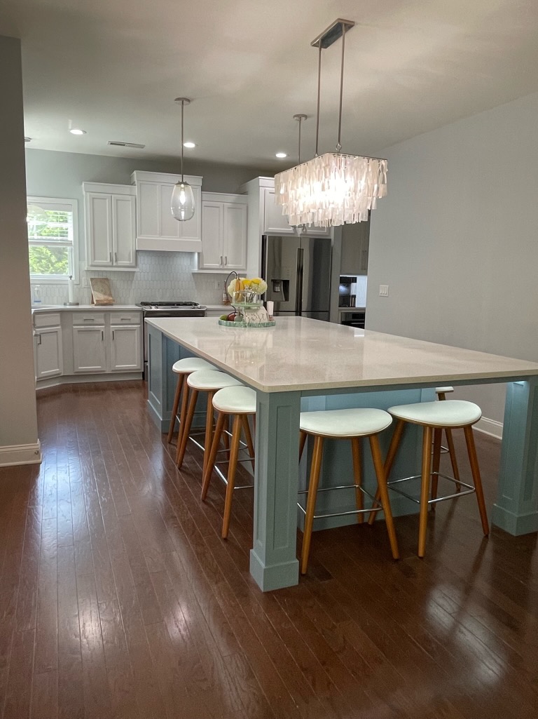 Modern kitchen with white cabinets, a large island featuring a teal base, four white bar stools, pendant and chandelier lighting, and dark wood flooring. A bowl of lemons decorates the island—showcasing top cabinet painting Charlotte style.