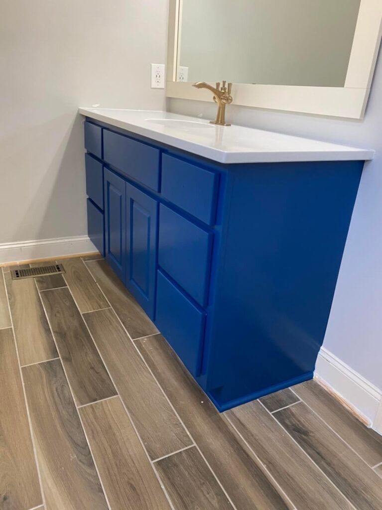 A modern bathroom vanity with bright blue cabinets, a white countertop, and a gold faucet. The wood-look tile floor completes the look—perfect inspiration for cabinet painting Charlotte homeowners will love. A large mirror hangs above the sink.