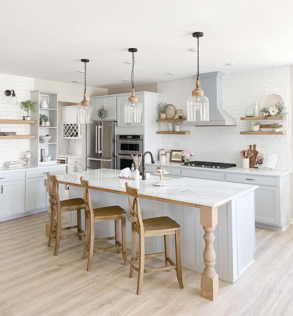 Bright, modern farmhouse kitchen with light wood floors, pale cabinets that showcase expert cabinet painting Charlotte style, marble island, wooden stools, open shelving, stainless steel appliances, and pendant lights. Decor includes plants and kitchenware.