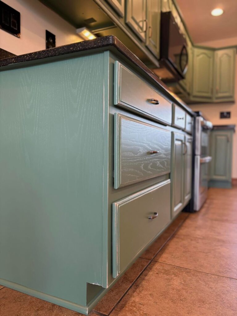 Close-up of lower kitchen cabinets painted light green with black handles, featuring a black countertop and tan tiled floor. A corner shows slight chipping at the base. Upper cabinets in the background highlight expert cabinet painting Charlotte services.
