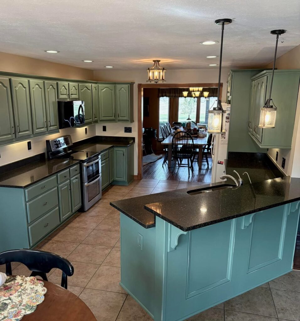 A kitchen with green cabinet painting Charlotte style, black countertops, stainless steel appliances, and pendant lights over a peninsula. Beyond the kitchen, a dining area with a large window and wooden furniture is visible.