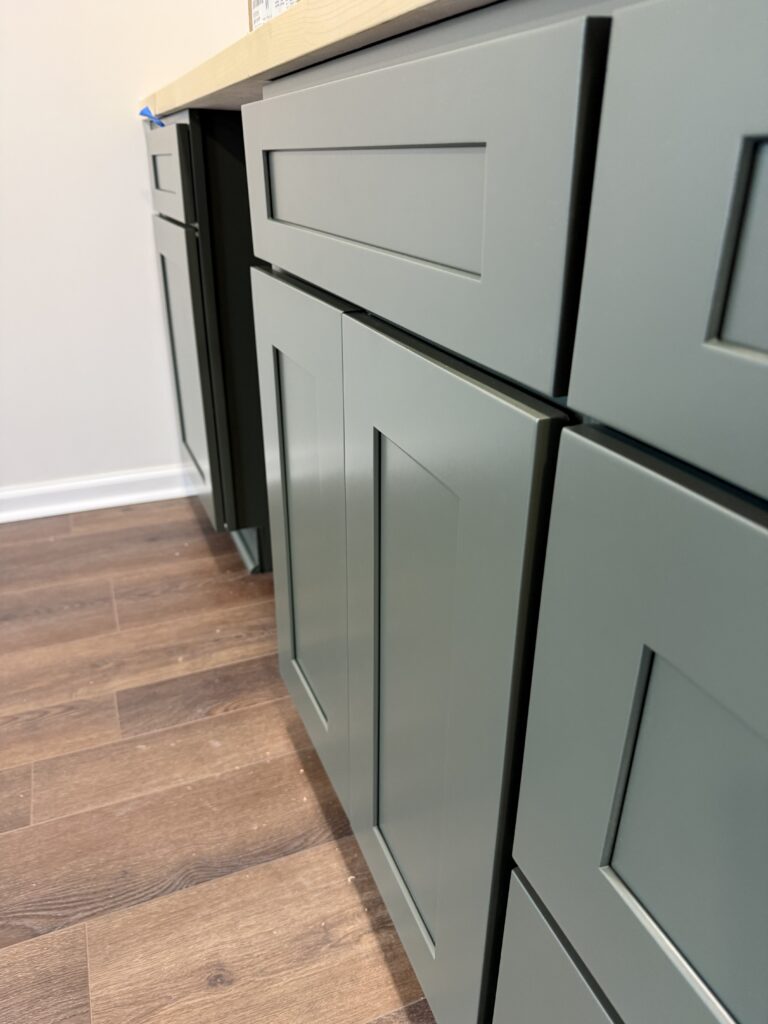 Close-up view of green shaker-style kitchen cabinets with silver handles, set against a wooden floor and a light-colored countertop—showcasing expert cabinet painting Charlotte homeowners love.
