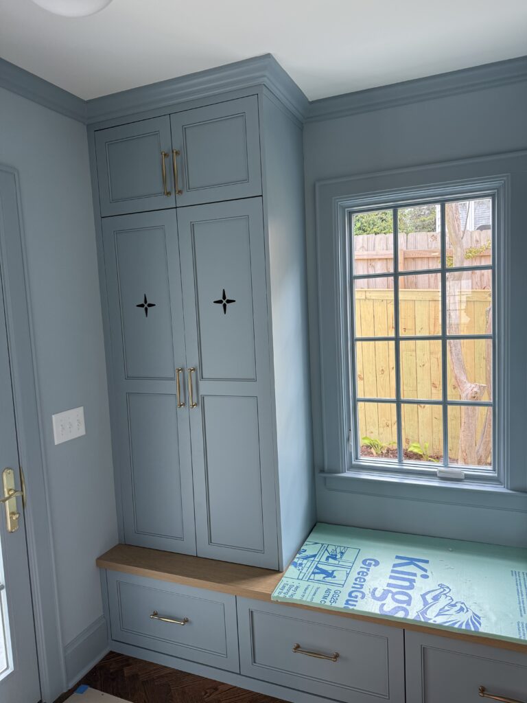 Light blue built-in cabinets with cross-shaped cutouts showcase expert cabinet painting Charlotte style, adjacent to a window seat with a wooden bench top. A package of bottled water sits on the bench beneath a window overlooking a fenced backyard.
