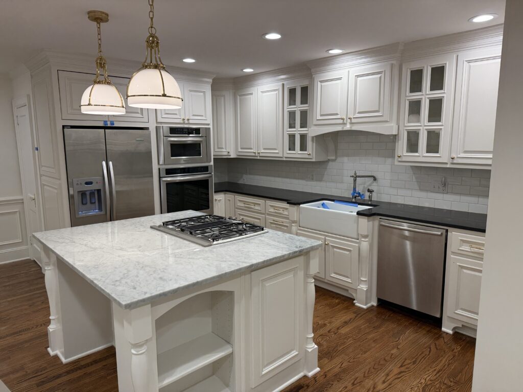 A modern kitchen featuring freshly done cabinet painting Charlotte, white cabinets, stainless steel appliances, a marble island with gas cooktop, farmhouse sink, two pendant lights, and gleaming hardwood floors.