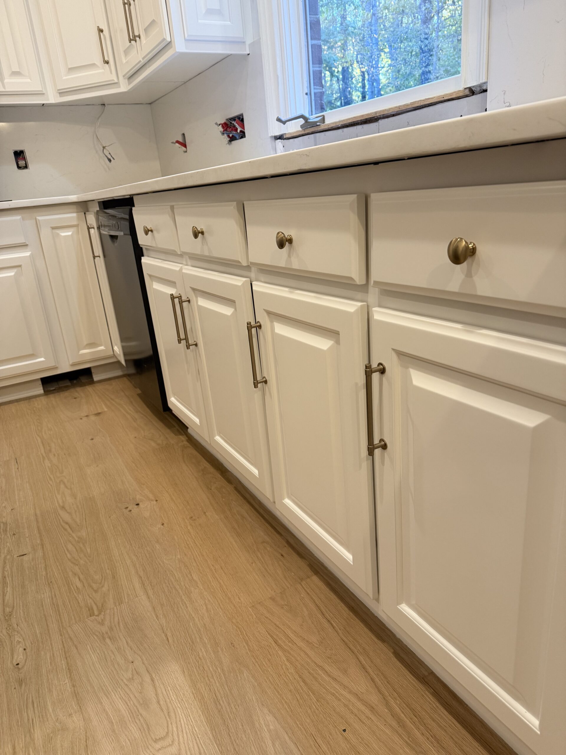 White kitchen cabinets with gold knobs and handles, finished by cabinet painting Charlotte experts, are installed beneath a white countertop. Wooden floors and a partially finished wall with exposed outlets are visible, along with a window above the countertop.