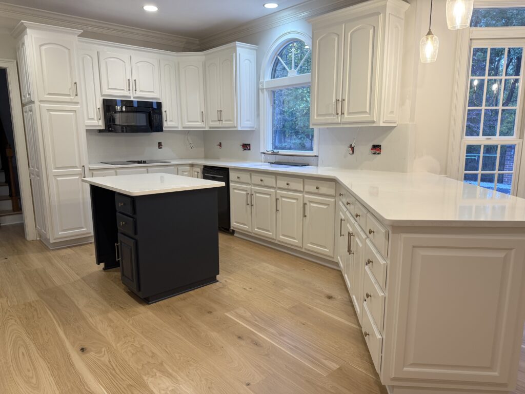 Modern kitchen with white cabinets, cabinet painting Charlotte expertise, light wood flooring, a center island with dark navy base, and quartz countertops. Large windows fill the space with natural light and recessed ceiling lights provide ample illumination.
