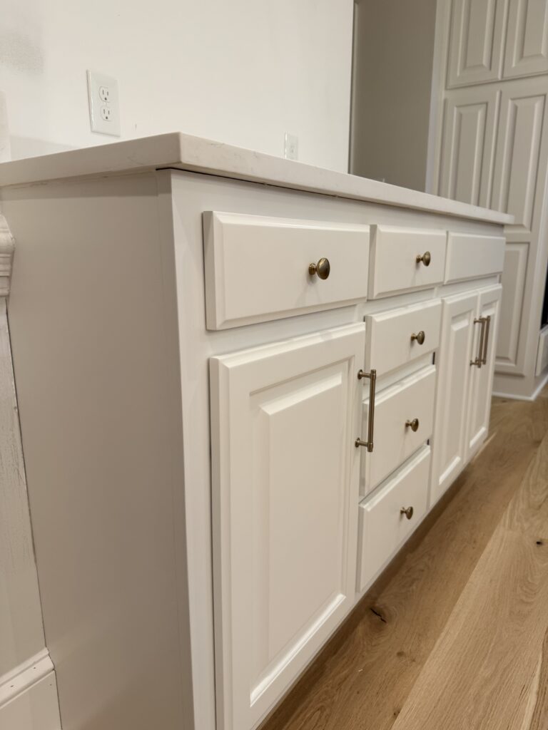 White bathroom vanity with gold hardware, marble countertop, and multiple drawers and cabinets—expert cabinet painting Charlotte services can help you achieve this fresh look. The vanity sits on light wood flooring, with a white paneled wall in the background.