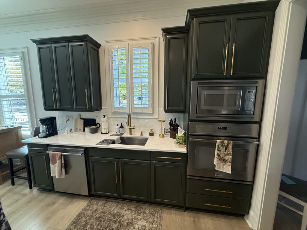 Modern kitchen with dark green cabinet painting charlotte finishes, gold handles, white countertops, stainless steel appliances, a coffee maker, and a sink below a window with white shutters. A towel hangs from the dishwasher.