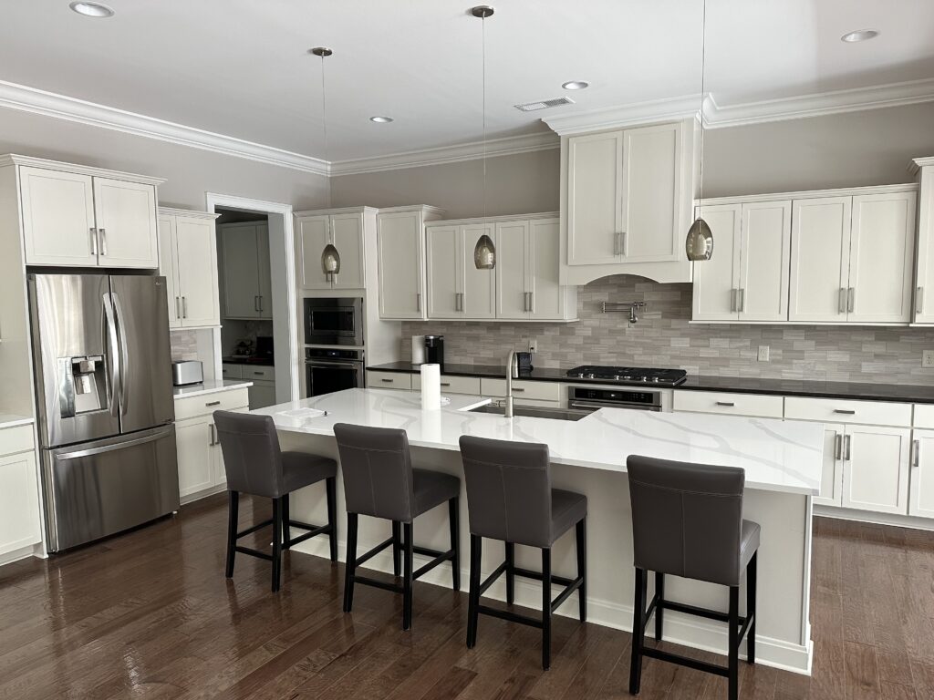 Modern kitchen with white cabinets, stainless steel appliances, a large island with four dark leather chairs, pendant lights above, and hardwood flooring. Expert cabinet painting Charlotte enhances the neutral tones and bright, open space.