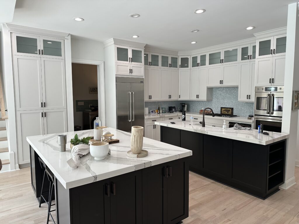 Modern kitchen with white upper cabinets, black lower cabinets, stainless steel appliances, and two marble islands. Decorative items are on the islands, and a mosaic tile backsplash accents this cabinet painting Charlotte dream space.