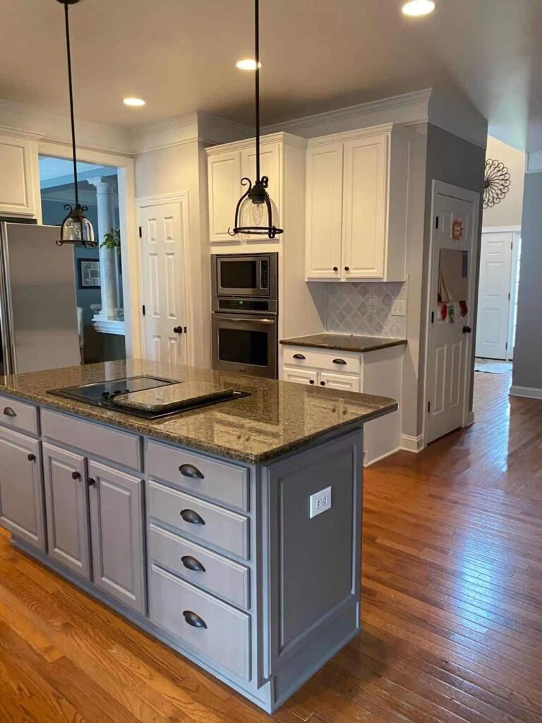 Modern kitchen with wooden floors, a large gray island with a built-in stovetop, white cabinets featuring expert cabinet painting Charlotte services, stainless steel appliances, pendant lighting, and neutral-colored walls.