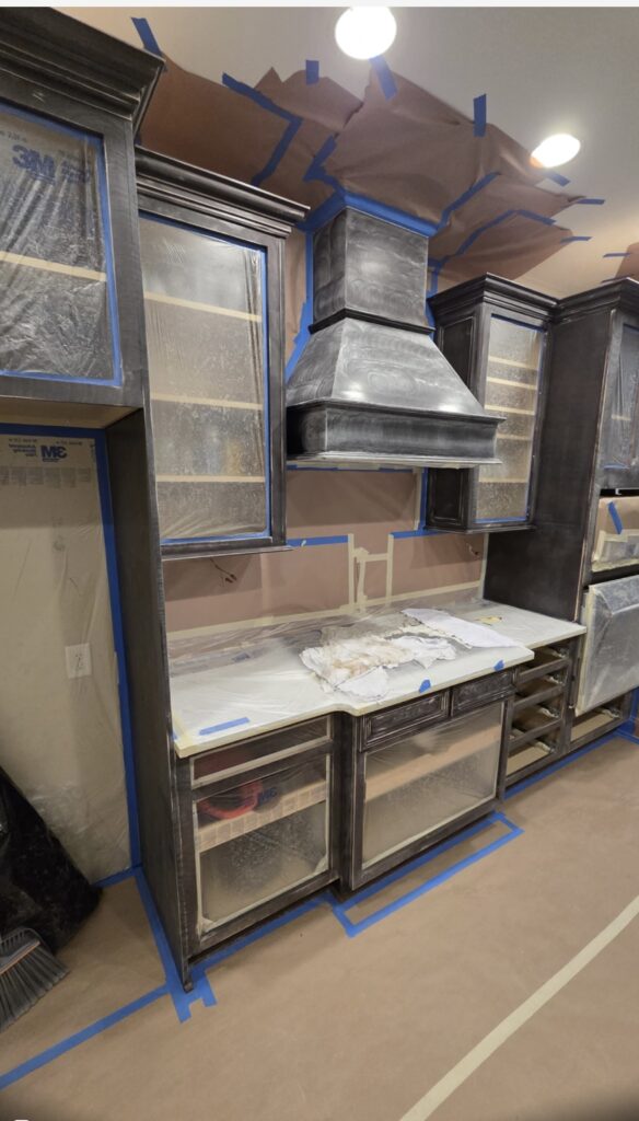 A kitchen under renovation with cabinets, range hood, and countertop covered in protective plastic and blue painter’s tape, ready for cabinet painting Charlotte. The floor and backsplash are protected, with tools visible on the counter.