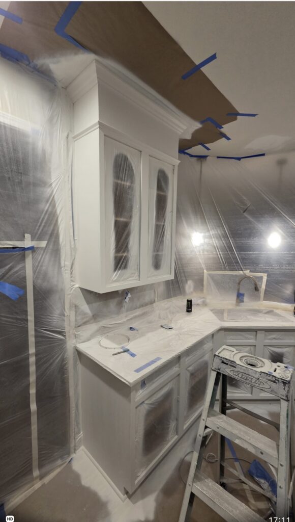 A kitchen under renovation, covered in plastic sheets and painter’s tape, shows cabinets and countertops primed white—a hallmark of cabinet painting Charlotte projects. A step ladder stands in front, with light fixtures visible through the plastic.