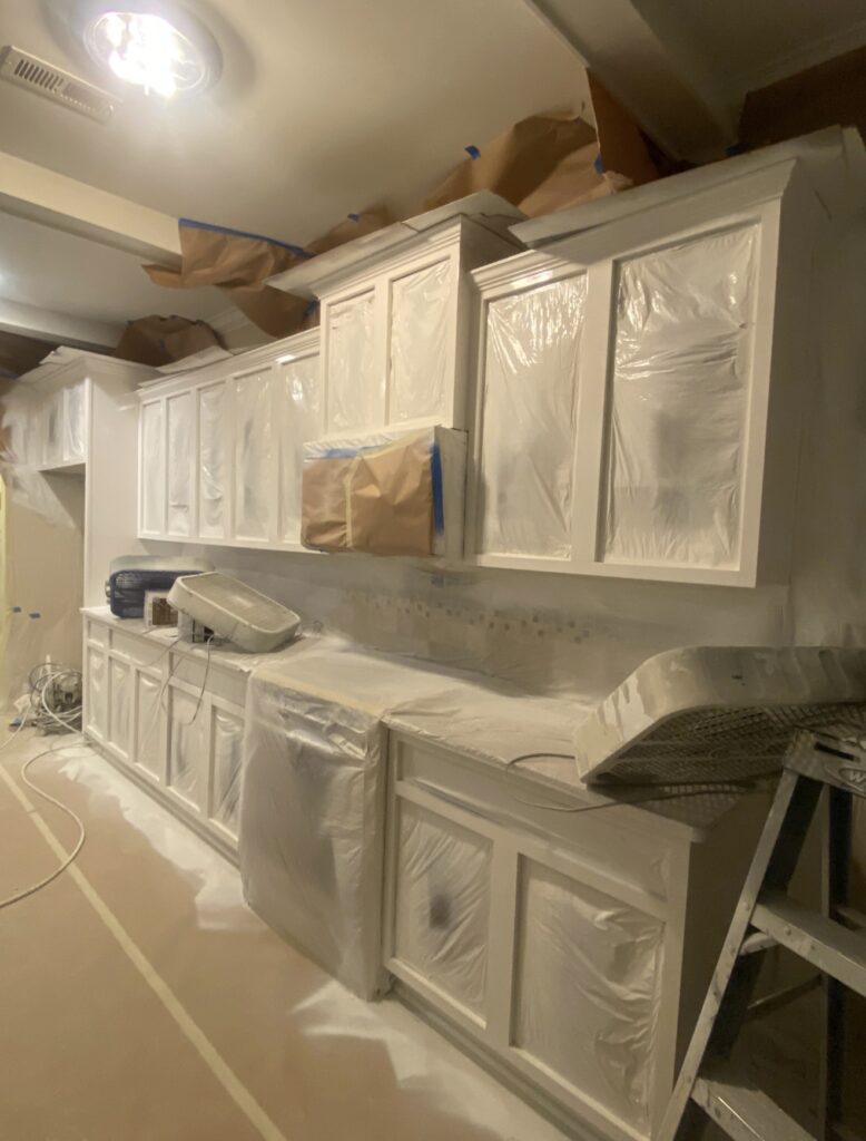 A kitchen prepared for cabinet painting Charlotte, with cabinets, counters, and appliances covered in plastic and paper. The floor is protected as well, and a ladder is visible on the right side, ready for renovation work.