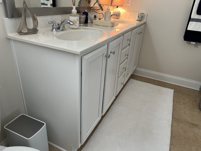 White bathroom vanity with two sinks, silver faucets, and cabinets. Decorative items, soap dispenser, tissue box, and a small lamp are on the counter. A white rug lies on the beige tile floor. Towels hang on the wall in the background.