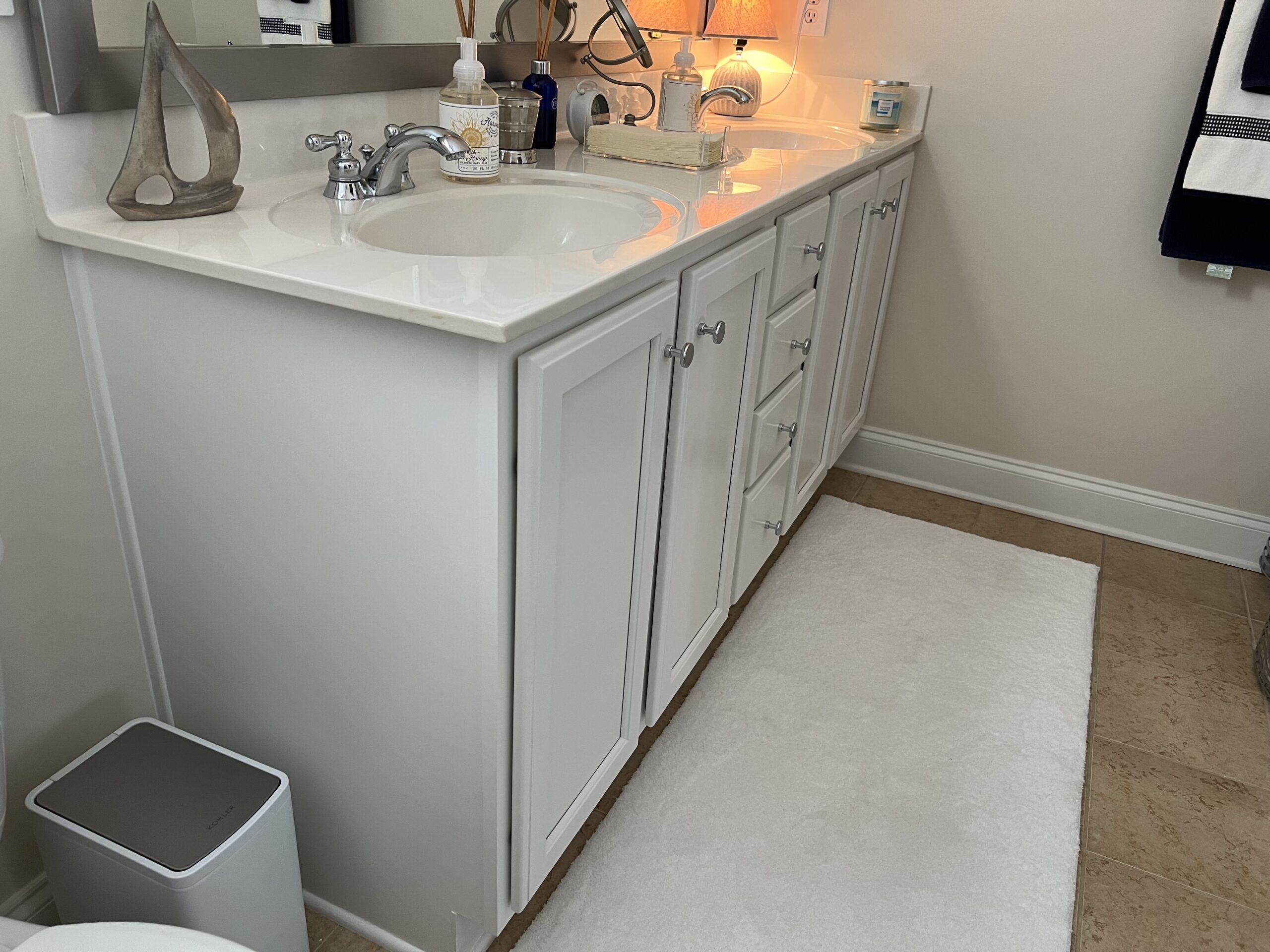 White bathroom vanity with two sinks, silver faucets, and cabinets. Decorative items, soap dispenser, tissue box, and a small lamp are on the counter. A white rug lies on the beige tile floor. Towels hang on the wall in the background.