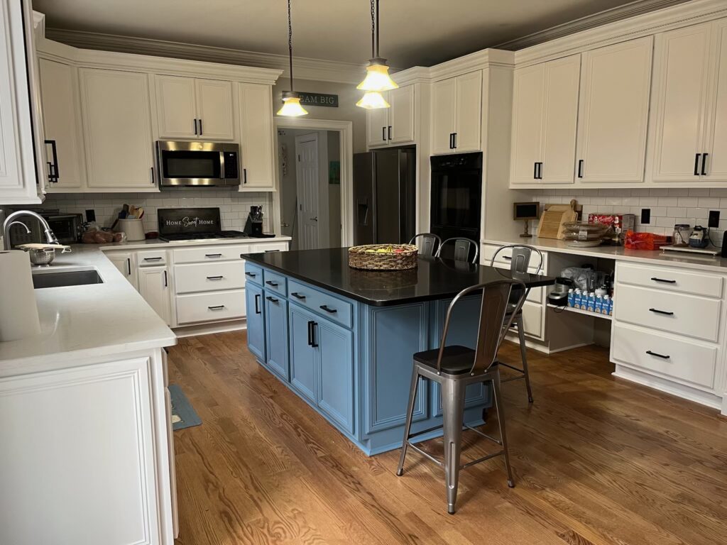 A spacious kitchen with white cabinets, stainless steel appliances, wooden floor, and a large blue island with a black countertop. Two metal chairs are at the island, showcasing the quality of cabinet painting Charlotte homeowners love.