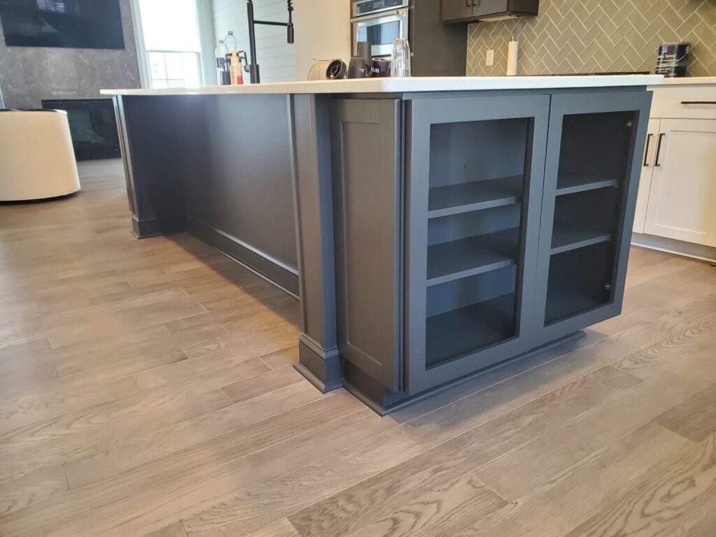 A modern kitchen island with dark blue cabinetry, glass-front lower cabinets, and a light countertop, set on light wood flooring—perfect inspiration for those considering cabinet painting Charlotte style.