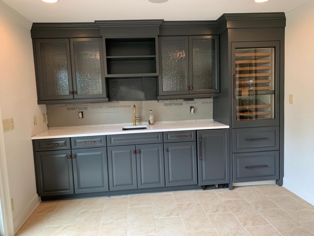 A modern kitchen with dark gray cabinets, glass upper cabinet doors, a built-in wine fridge, a white countertop, and beige tile flooring. Perfect for those considering cabinet painting Charlotte for a fresh update; the backsplash area is unfinished.