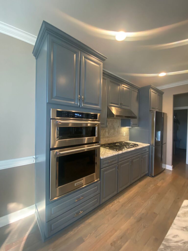 Modern kitchen with gray cabinets, stainless steel double oven, gas cooktop, vent hood, and refrigerator. Light wood flooring and neutral walls reflect expert cabinet painting Charlotte style, with crown molding and recessed lighting overhead.
