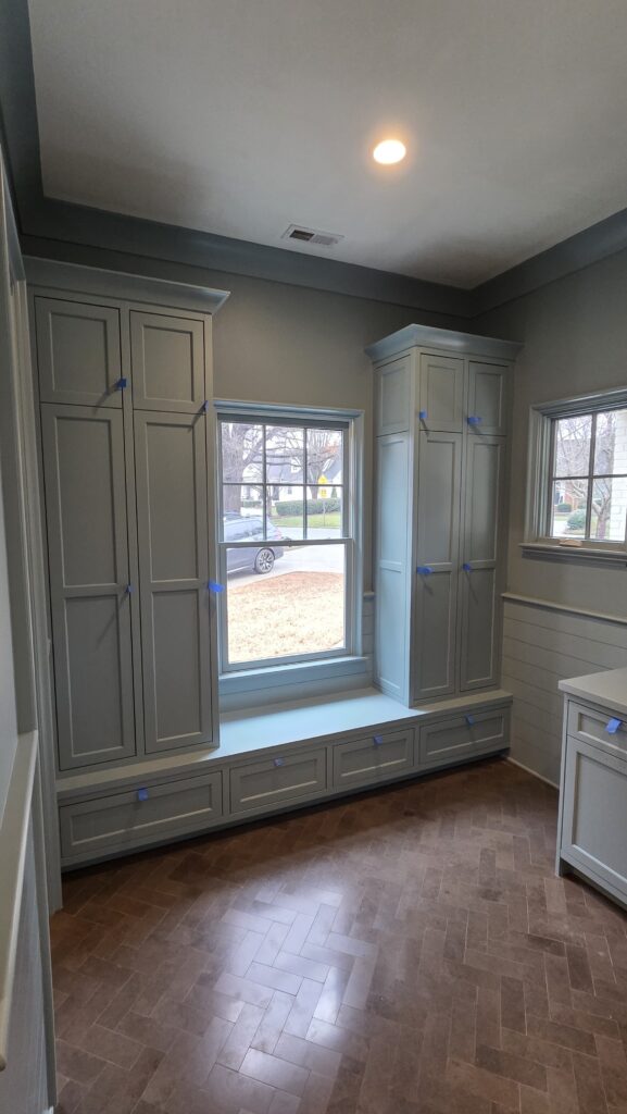A room with light gray built-in cabinets, freshly marked for cabinet painting Charlotte. A window seat rests beneath two large windows, while blue painter’s tape outlines cabinet edges. The herringbone-pattern wood floor completes the stylish look. A car is visible outside.