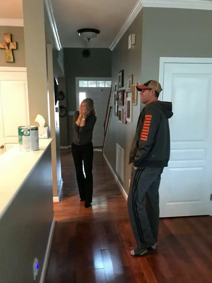 A woman stands in a hallway covering her mouth with her hands, while a man in a cap, hoodie, and sweatpants stands nearby. They are in a home with wooden floors and gray walls decorated with photos.