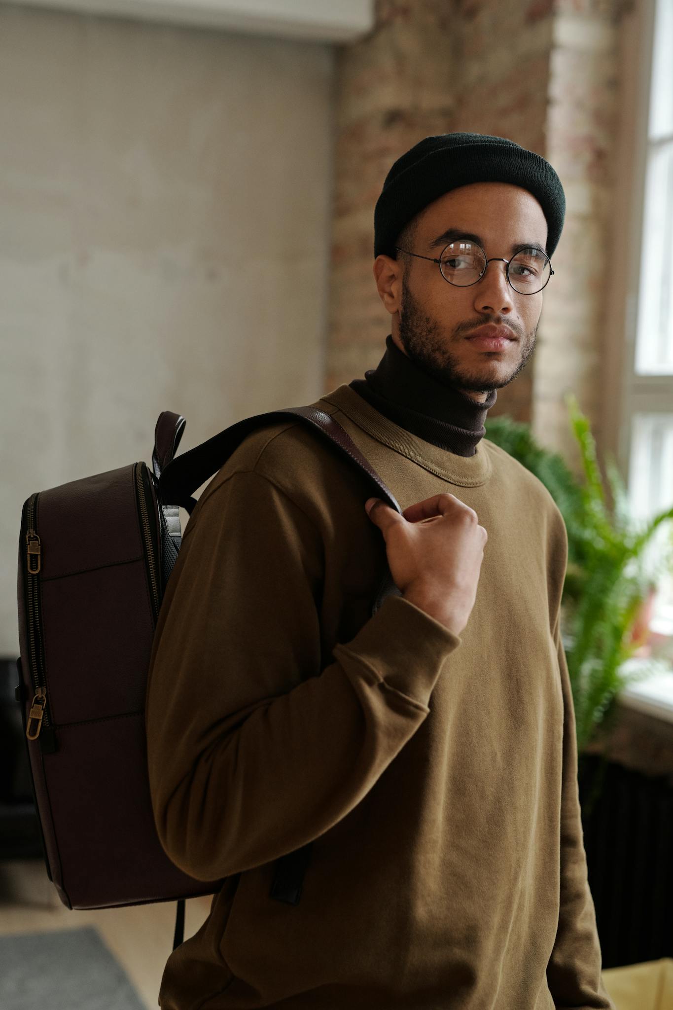 A young man wearing glasses, a black beanie, and a brown sweatshirt stands indoors with a backpack over one shoulder, looking at the camera. A window, green plant, and a sign for cabinet painting Charlotte are in the background.