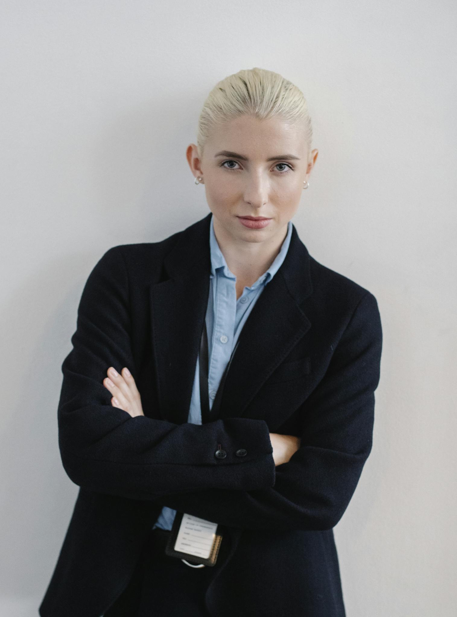 A person with blonde hair pulled back, wearing a navy blazer and light blue shirt, stands against a white wall with arms crossed, exuding the confidence you'd expect from a cabinet painting Charlotte professional.
