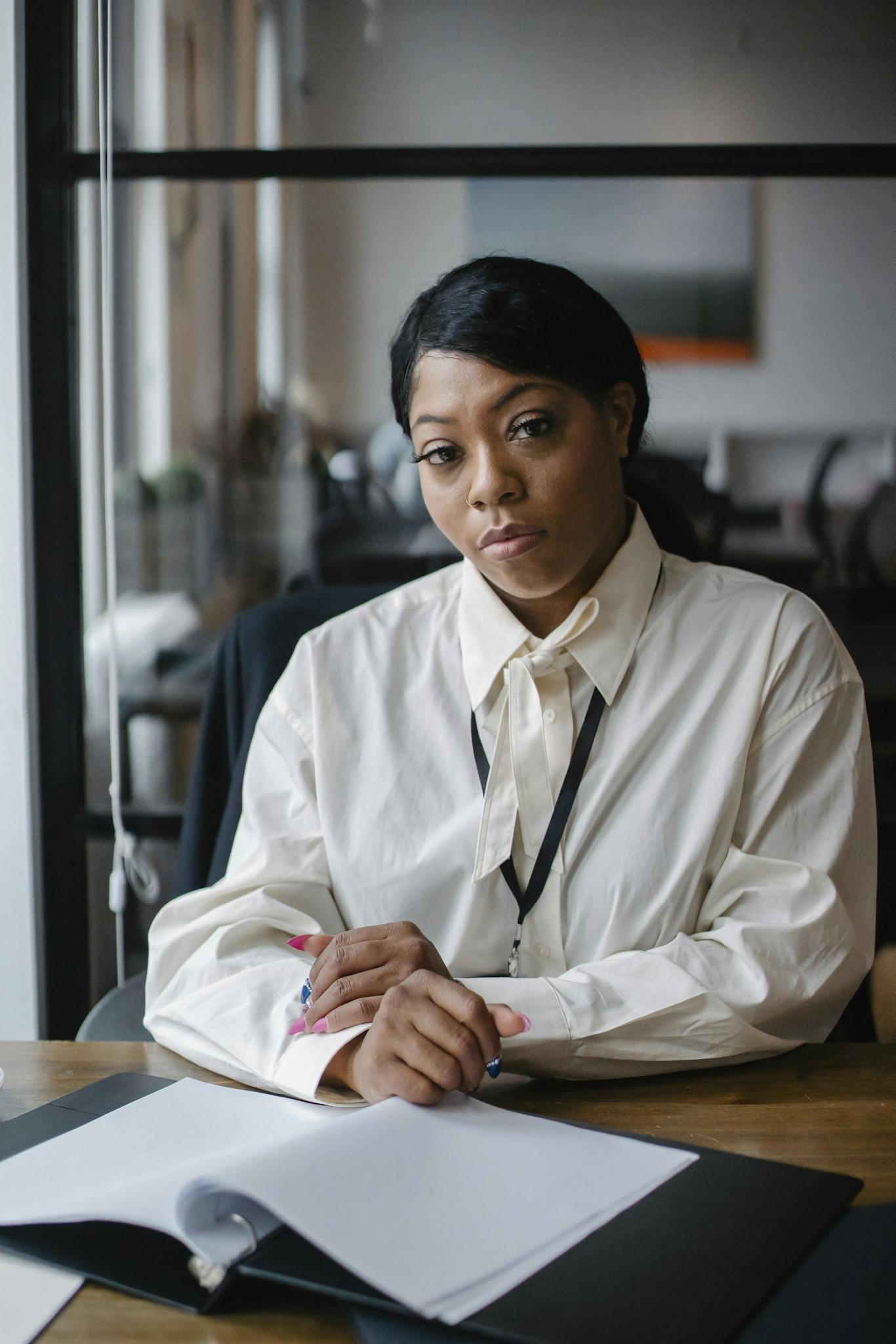 A woman wearing a white blouse and lanyard sits at a desk with an open notebook in front of her, looking at the camera with a serious expression—perhaps planning her next cabinet painting Charlotte project.