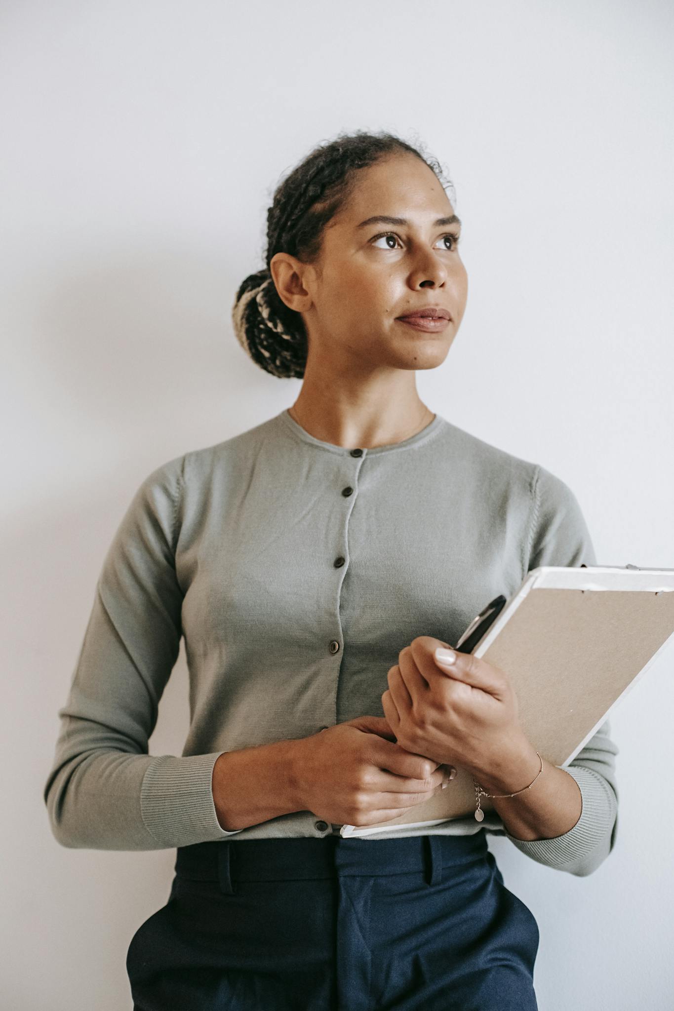 A woman wearing a light green cardigan and dark pants holds a clipboard and pen, looking thoughtfully to the side—perhaps considering a cabinet painting Charlotte project—while standing against a plain white background.