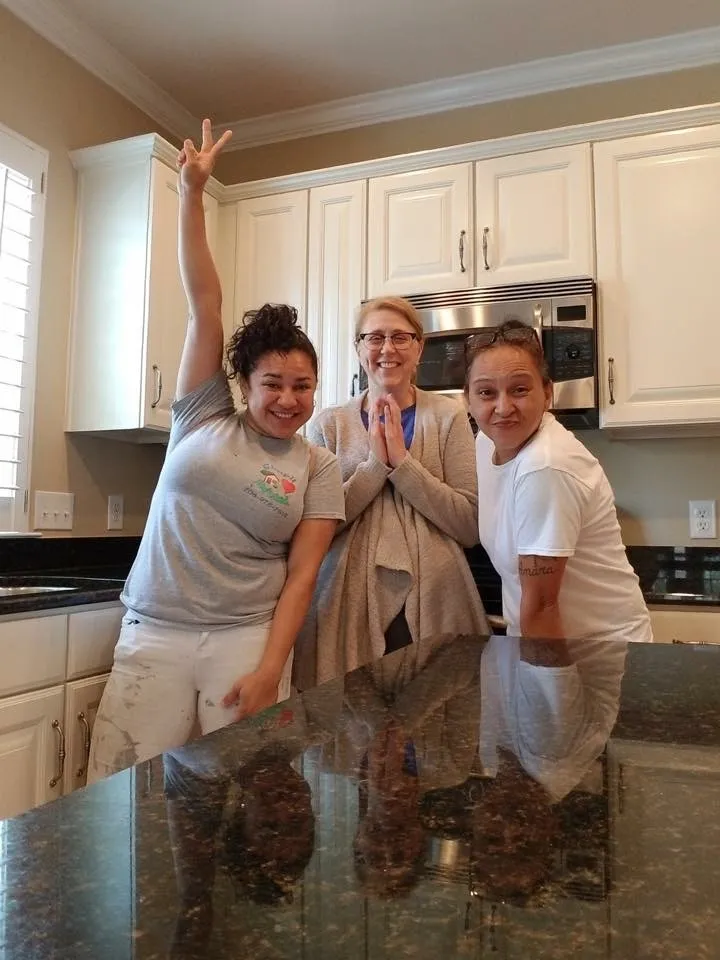 Three women smile in a kitchen with white cabinets; one on the left raises her hand in a peace sign, the middle woman clasps her hands, and the right leans forward, all happily posing by a granite countertop.