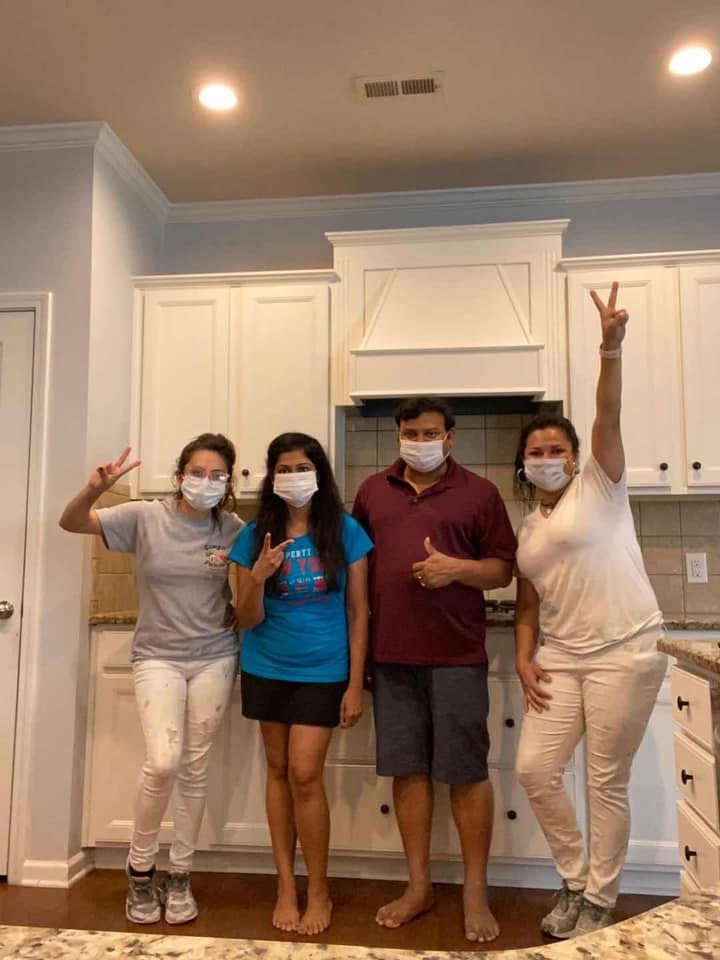 Four people wearing face masks stand in a kitchen, posing for a photo. Three of them make peace signs with their hands, while one gives a thumbs up. They appear happy and relaxed.