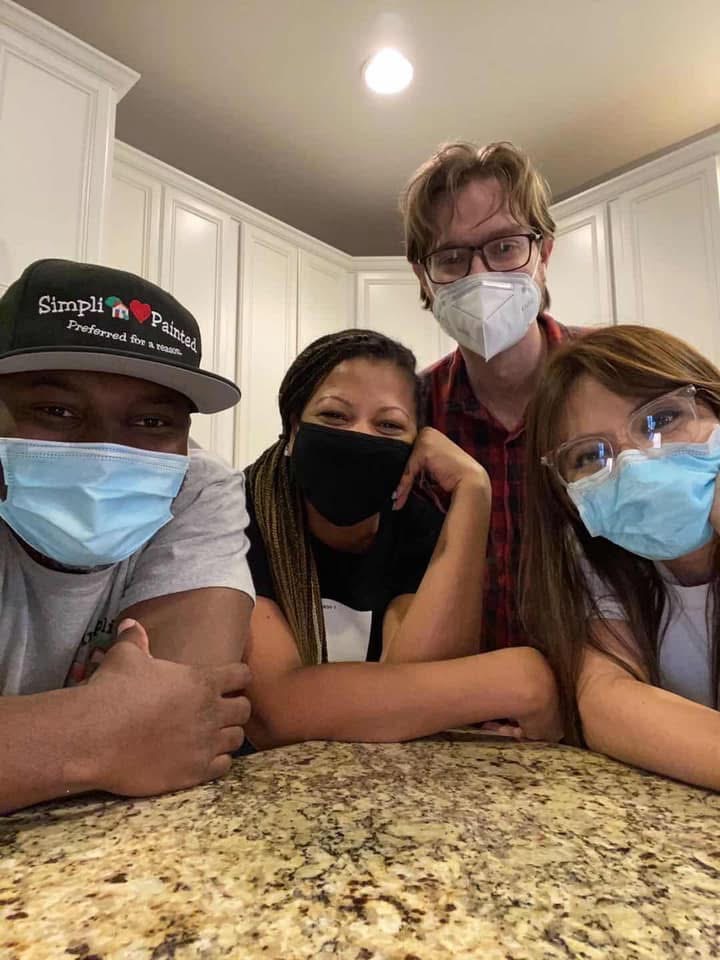 Four people wearing face masks and glasses are smiling and leaning on a kitchen counter, posing for a group photo in a brightly lit kitchen with white cabinets.
