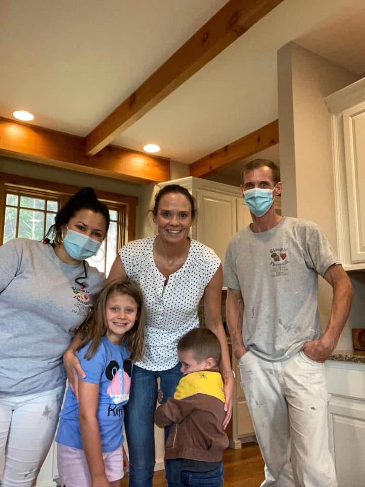 Four adults (two wearing masks) and two young children stand together in a kitchen with wooden beams, smiling at the camera. One child hugs an adults leg, while the others pose for the photo.