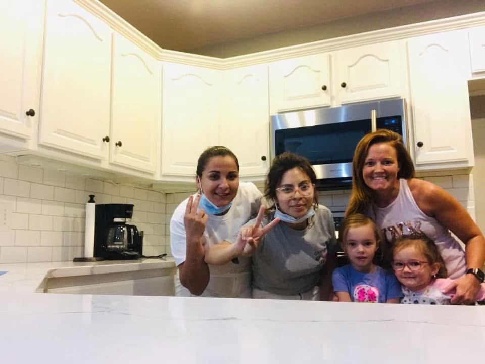 Five people, including two women in casual clothes, two young girls, and another young woman, pose and smile together in a bright kitchen with white cabinets and a microwave in the background.