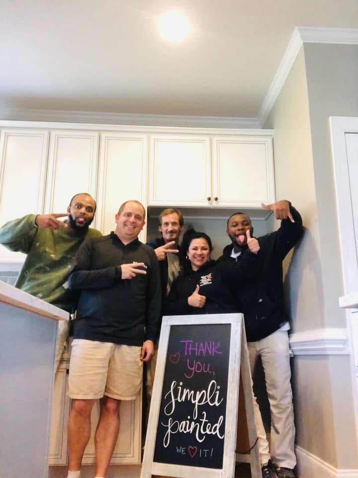 Five people smiling and posing together in a kitchen behind a sign that says, “Thank you, Simpli Painted, we ♥ it!” The atmosphere is cheerful, and the group appears to be celebrating the completed paint job.