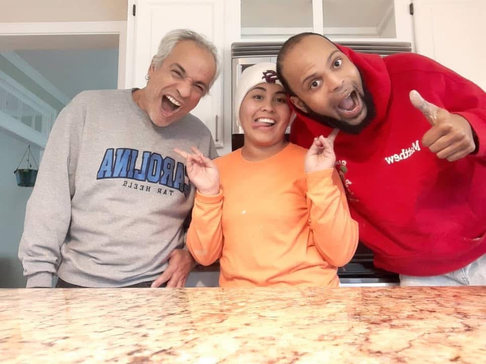 Three adults stand in a kitchen, smiling and making playful faces. The person on the left wears a gray sweatshirt, the center person an orange shirt and beanie, and the right person a red hoodie, giving a thumbs up.