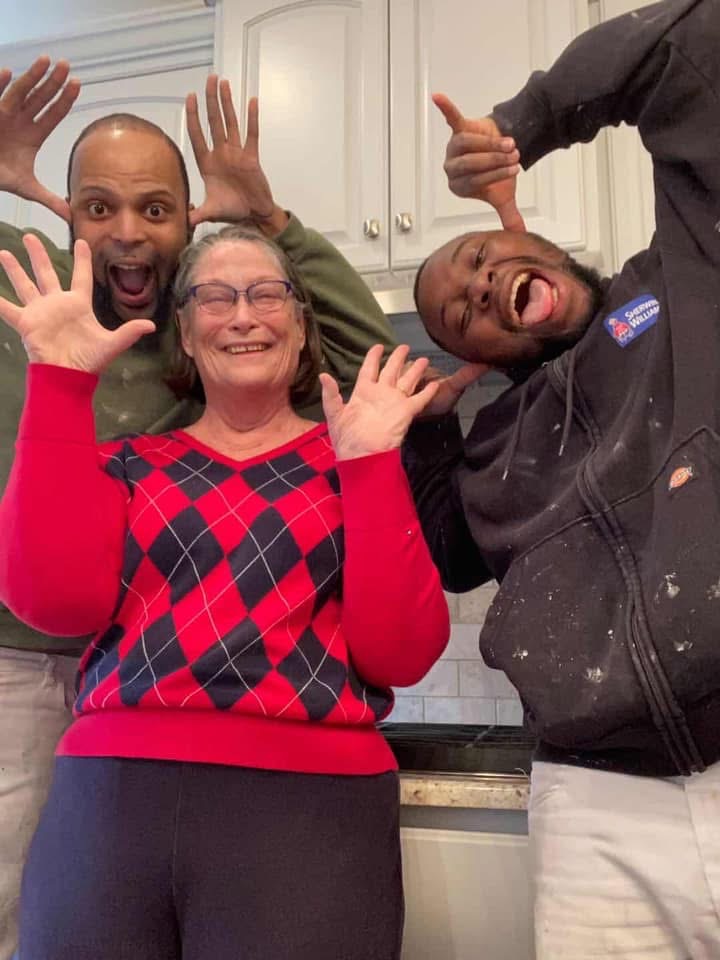 Three people stand in a kitchen smiling and making playful hand gestures toward the camera. The woman in the center wears glasses and a red argyle sweater, while the two men beside her are dressed casually and look cheerful.