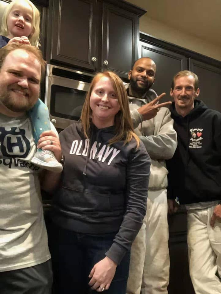 Five adults and one young child smiling and posing together in a kitchen, with dark cabinets visible in the background. One man is holding the child on his shoulder, and another man is making a peace sign.